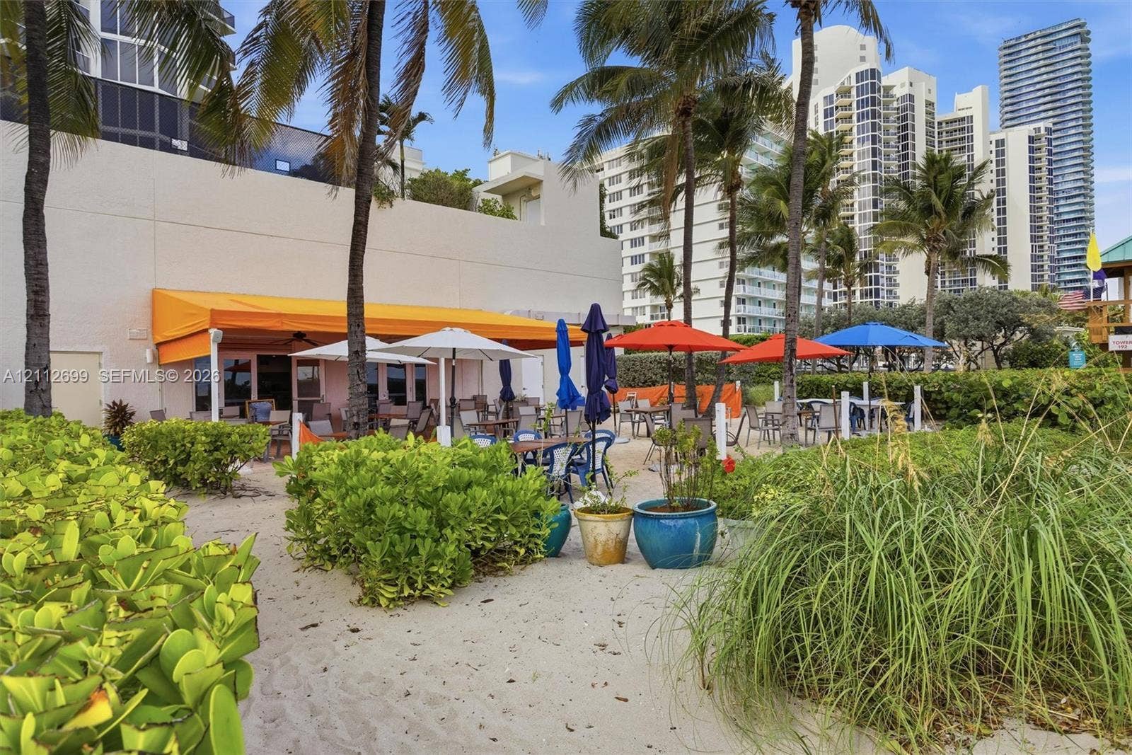 19111 Collins Avenue, #505, Sunny Isles Beach, FL 33160 Outdoor patio area featuring a retractable orange awning, sandy ground surface, mature palm trees, and native coastal vegetation