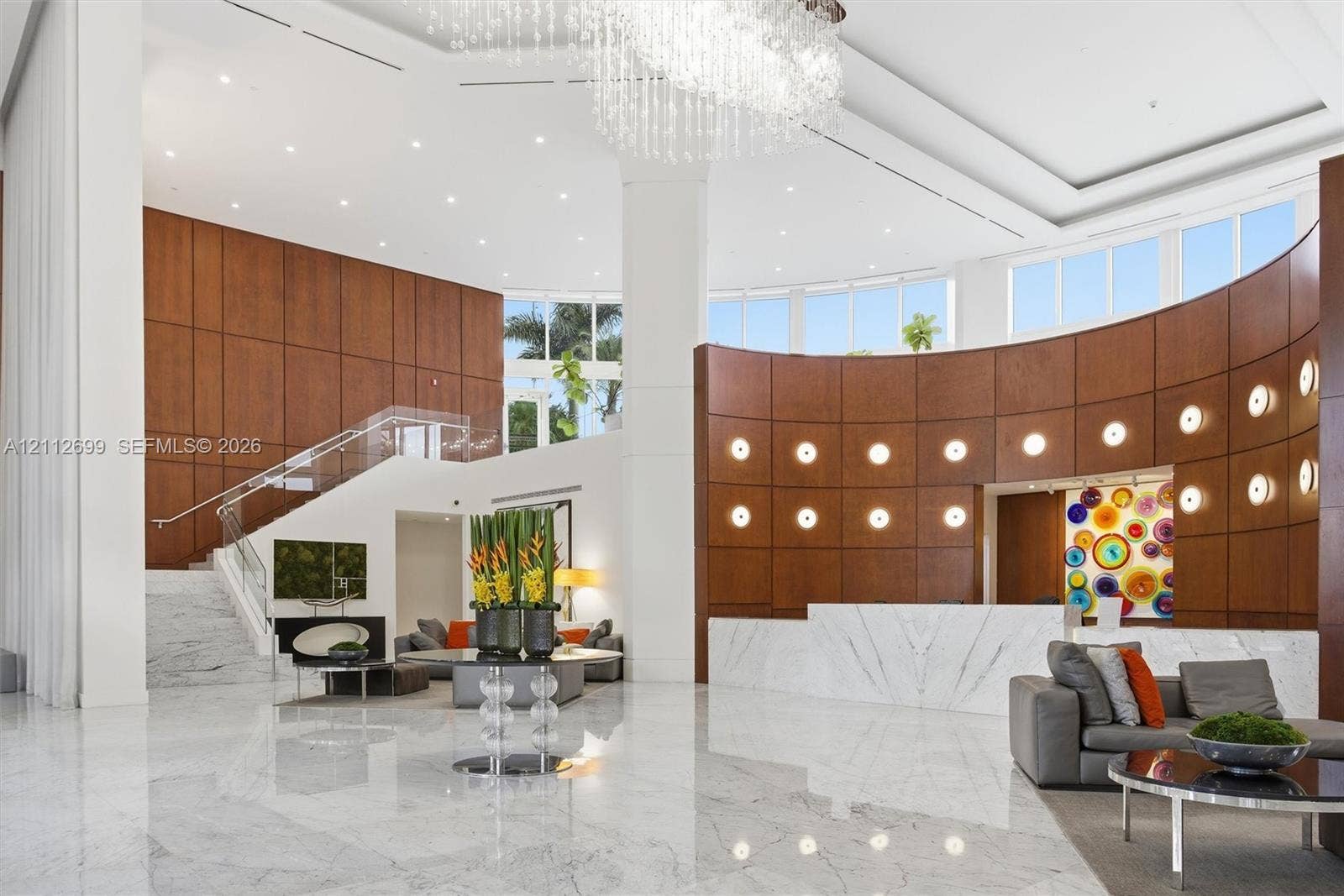 19111 Collins Avenue, #505, Sunny Isles Beach, FL 33160 Grand foyer featuring floor-to-ceiling wood paneling, curved architectural walls, polished stone flooring, a multi-tiered crystal chandelier, and a staircase with glass railings