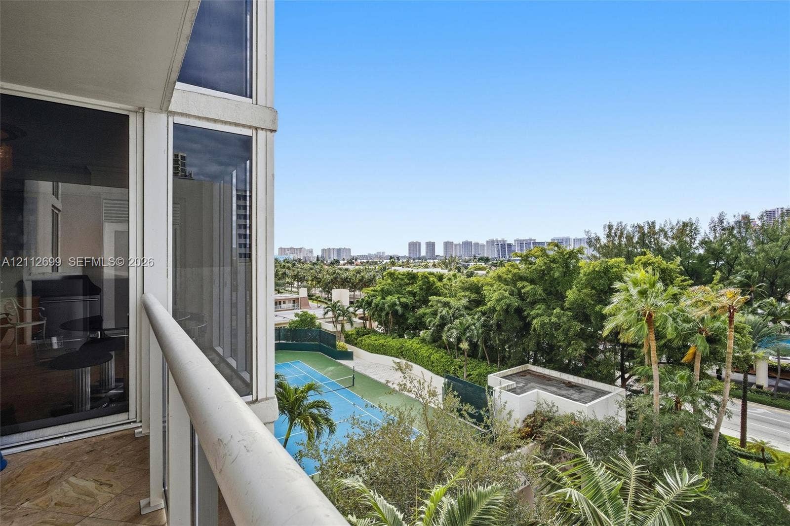 19111 Collins Avenue, #505, Sunny Isles Beach, FL 33160 Private balcony featuring stone-finish flooring and a glass-paneled railing, overlooking a tennis court and established palm trees