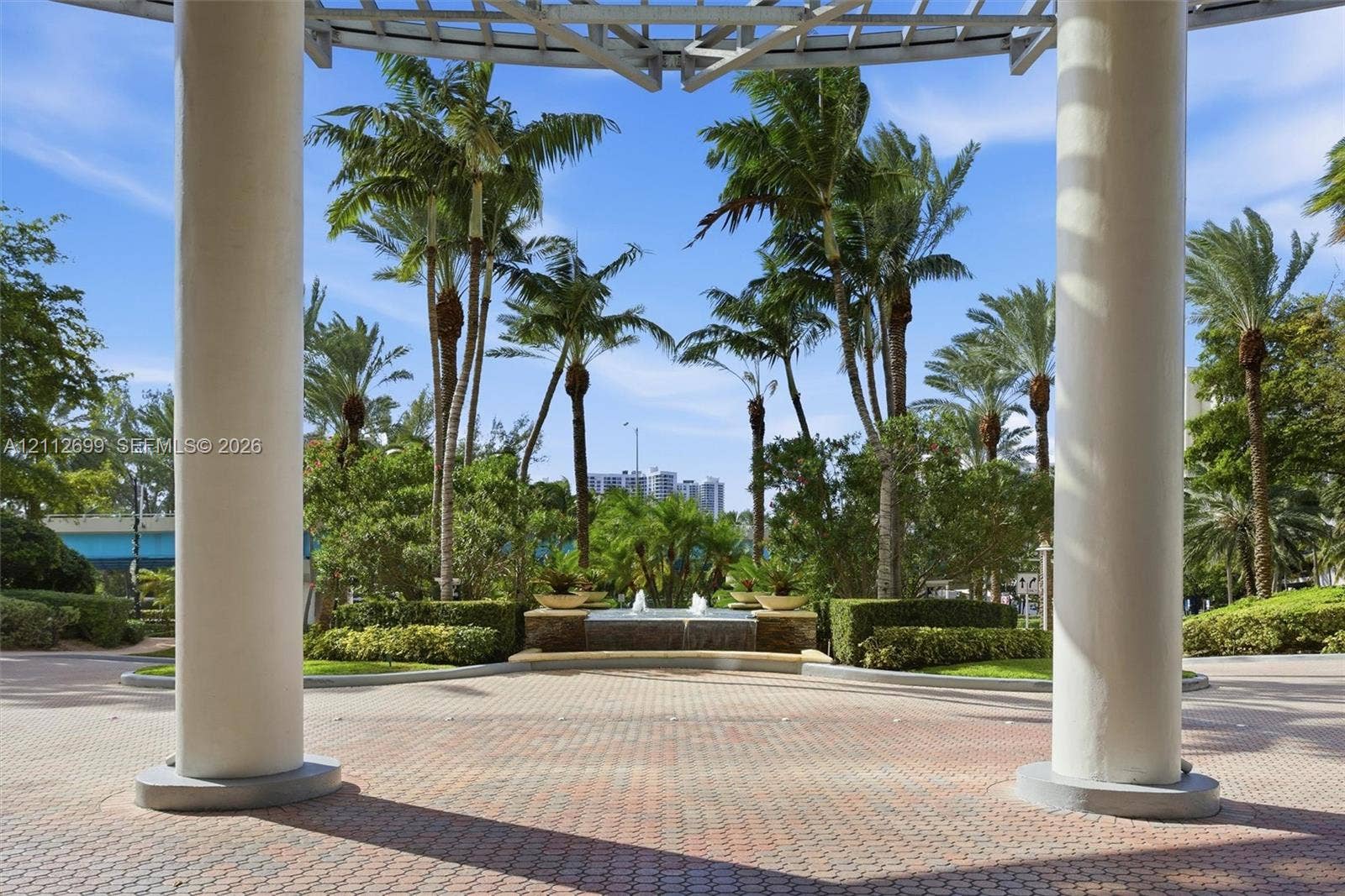 19111 Collins Avenue, #505, Sunny Isles Beach, FL 33160 Covered entrance featuring structural columns and a paver stone patio, centered fountain, and mature palm landscaping
