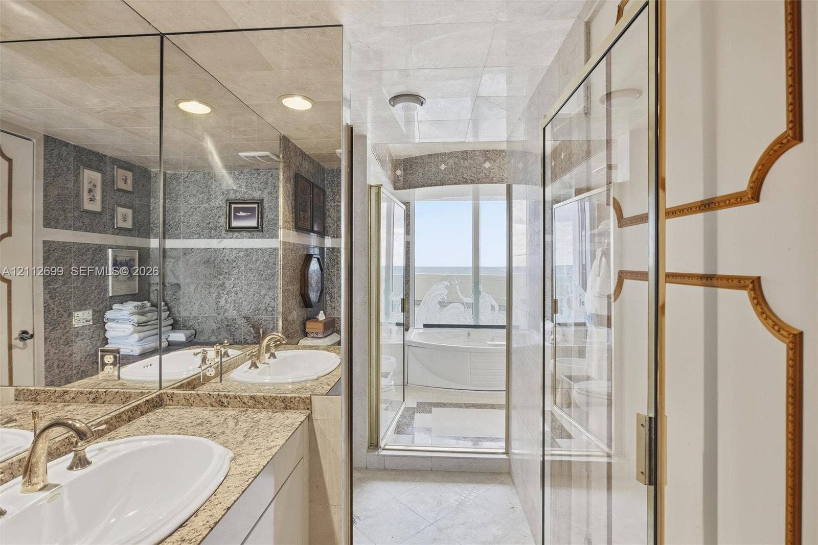 19111 Collins Avenue, #505, Sunny Isles Beach, FL 33160 Bathroom featuring a stone-topped dual vanity with integrated sinks, floor-to-ceiling mirrors, tile wall detailing, a jetted soaking tub, and custom paneled doors with gold-tone trim