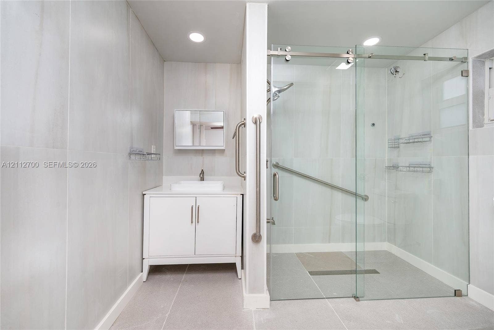 Bathroom featuring large-format wall tile, white vanity cabinet with a rectangular vessel sink, walk-in shower with glass sliding doors, and brushed nickel hardware