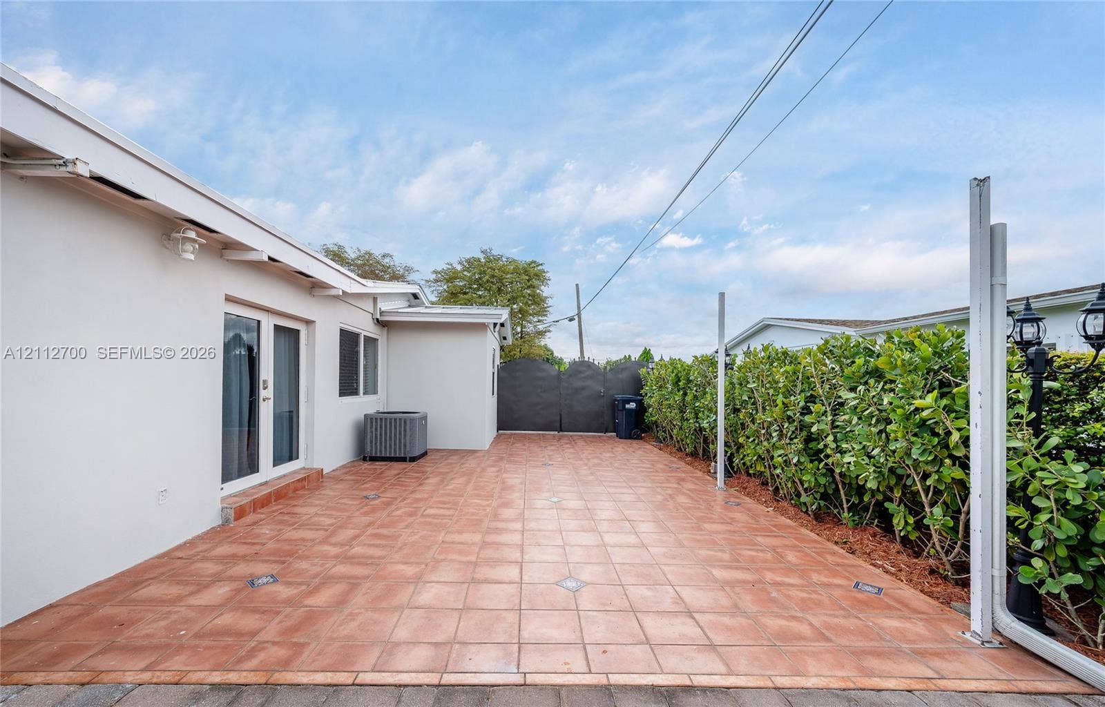 Rear exterior featuring a tiled patio, masonry privacy fencing, mature hedge landscaping, and wall-mounted exterior lighting