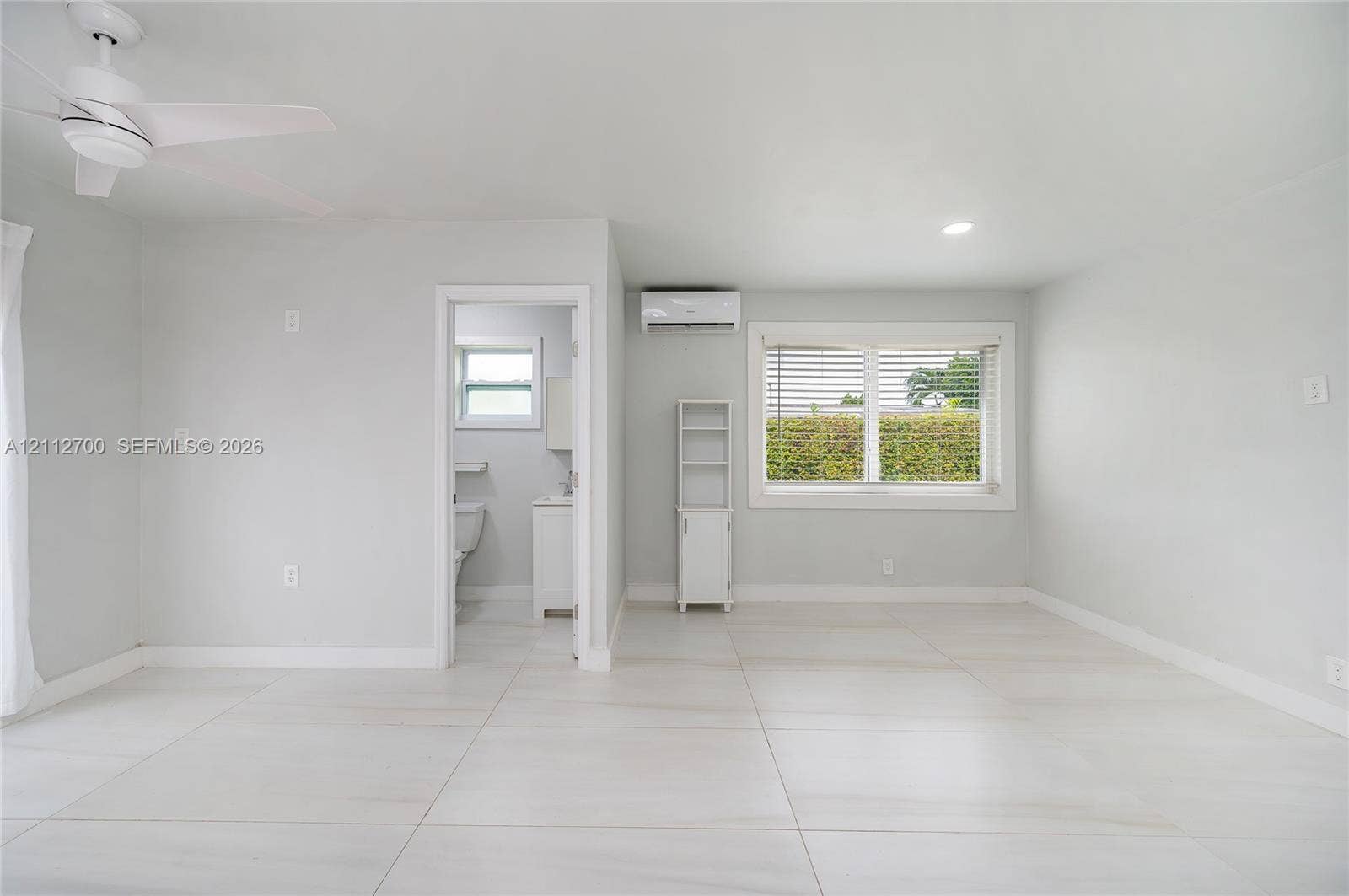 Large-format tile flooring, ceiling fan with integrated lighting, wall-mounted ductless air conditioning unit, and expansive window with horizontal blinds