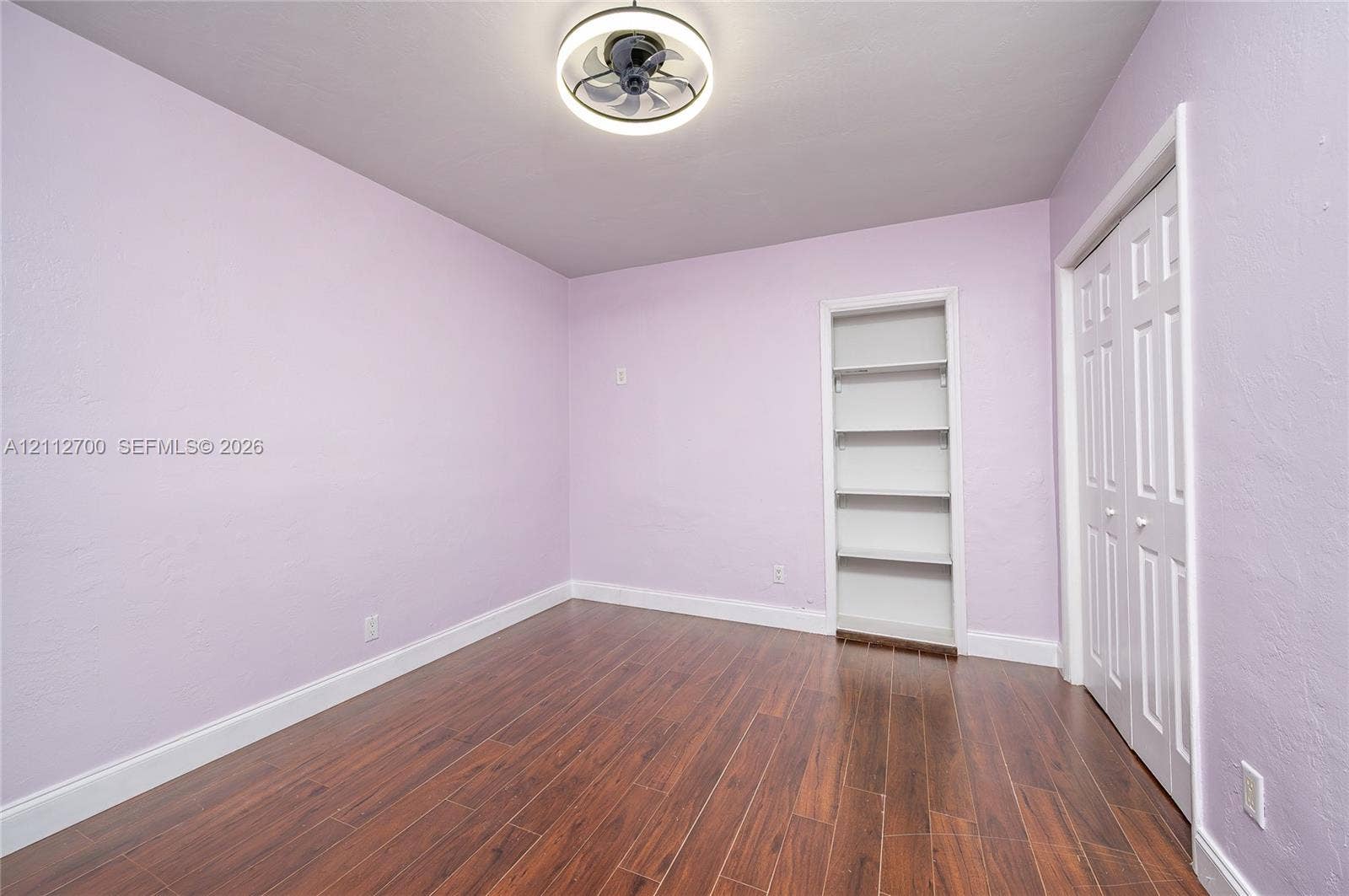 Interior space featuring wood-finish flooring, a built-in recessed wall shelving unit, double-door closet, and modern circular ceiling fan with integrated lighting