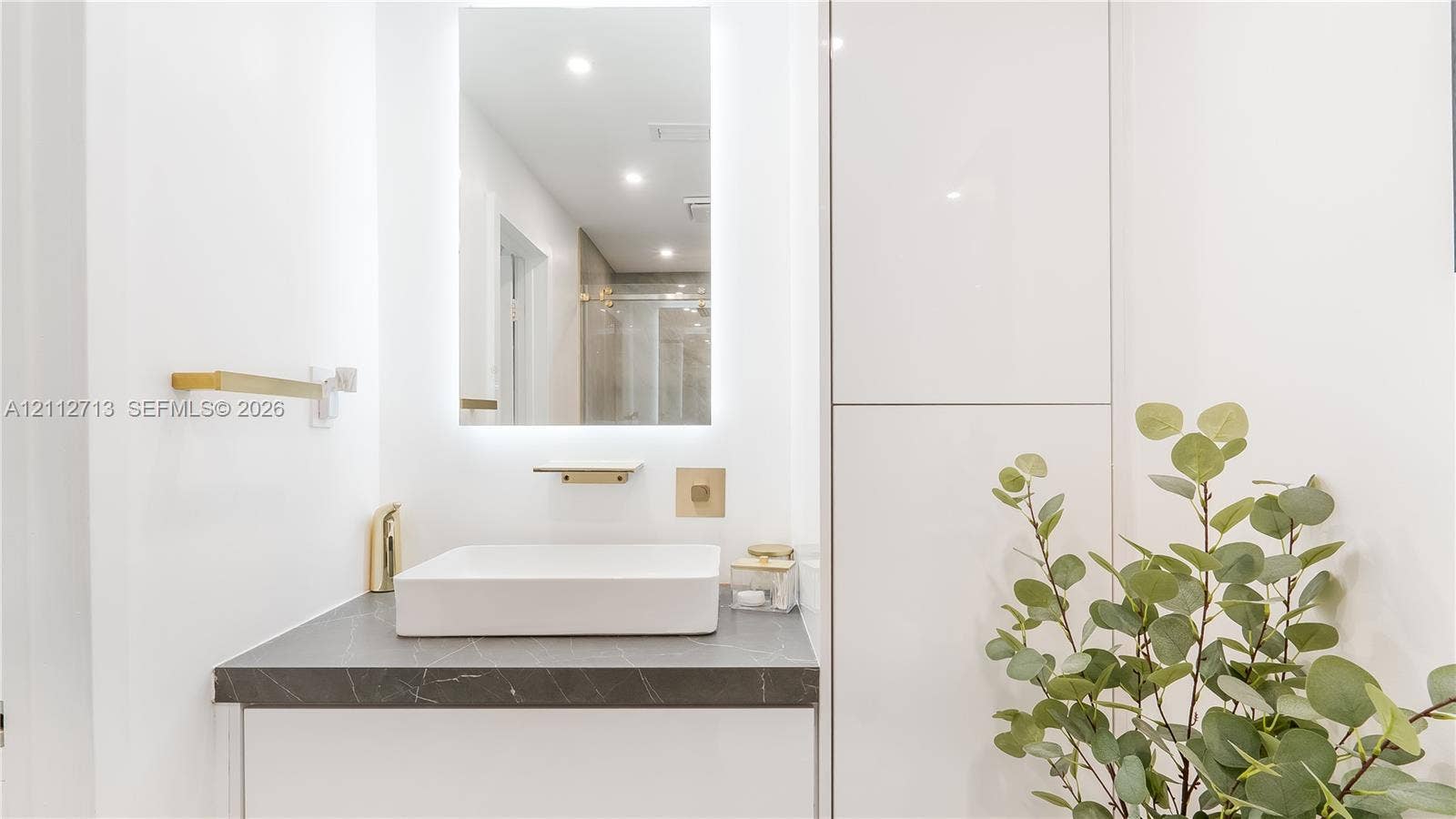 Modern vanity featuring a rectangular vessel sink, dark stone-finish countertop, integrated tall cabinetry, gold-tone hardware, and an LED-backlit mirror
