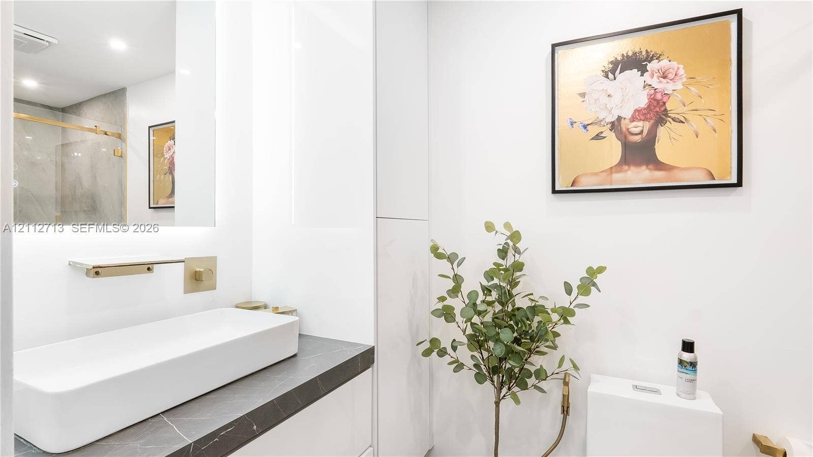 Modern vanity featuring a dark stone-finish countertop, white rectangular vessel sink, wall-mounted gold-tone faucet, and an illuminated mirror