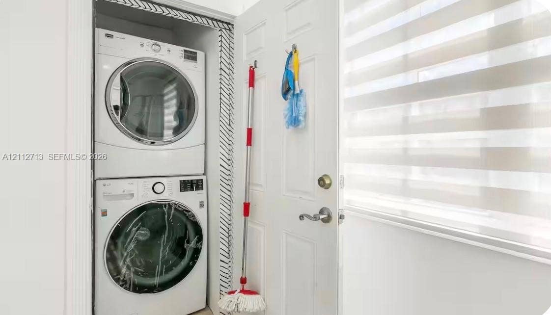Stacked laundry unit featuring a front-loading washer and dryer, housed within a closet featuring patterned interior trim and a multi-panel door with metallic hardware, accompanied by horizontally-striped roller blinds