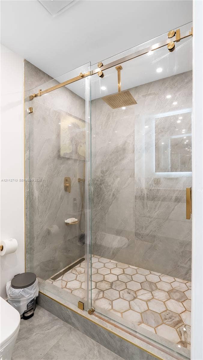 Walk-in shower featuring a frameless glass sliding door, metallic gold-tone hardware, a rainfall showerhead, large-format tile surround, and hexagonal tile flooring