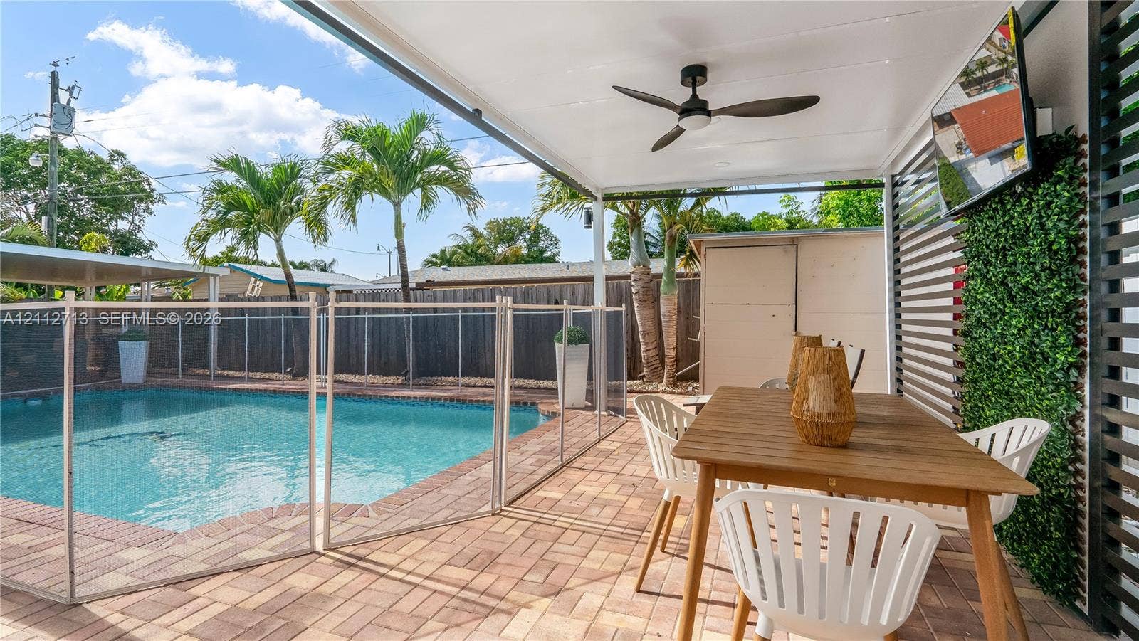 Covered patio featuring a ceiling fan, paver flooring, horizontal slat wall, and attached vertical garden wall, leading to a swimming pool with a safety enclosure