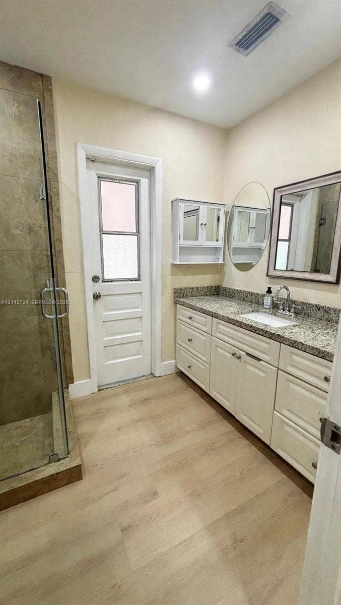 Walk-in shower with tiled surround, light-finish cabinetry with stone-style countertop, oval wall mirror, and wood-finish flooring