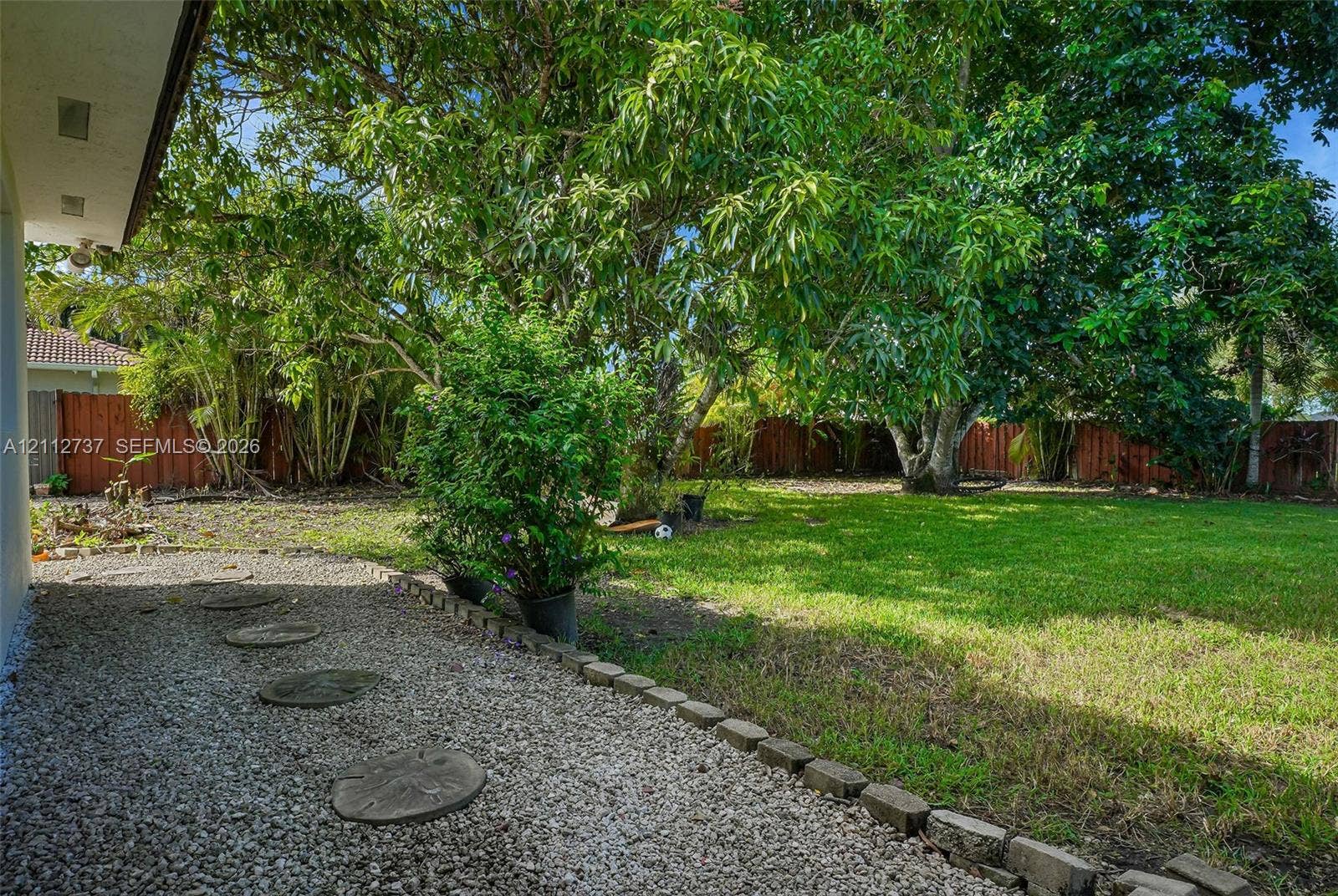 27902 SW 159th Court, Homestead, FL 33031 Private backyard featuring a manicured lawn, stone-border edging, gravel pathway with round pavers, and mature trees enclosed by wood fencing