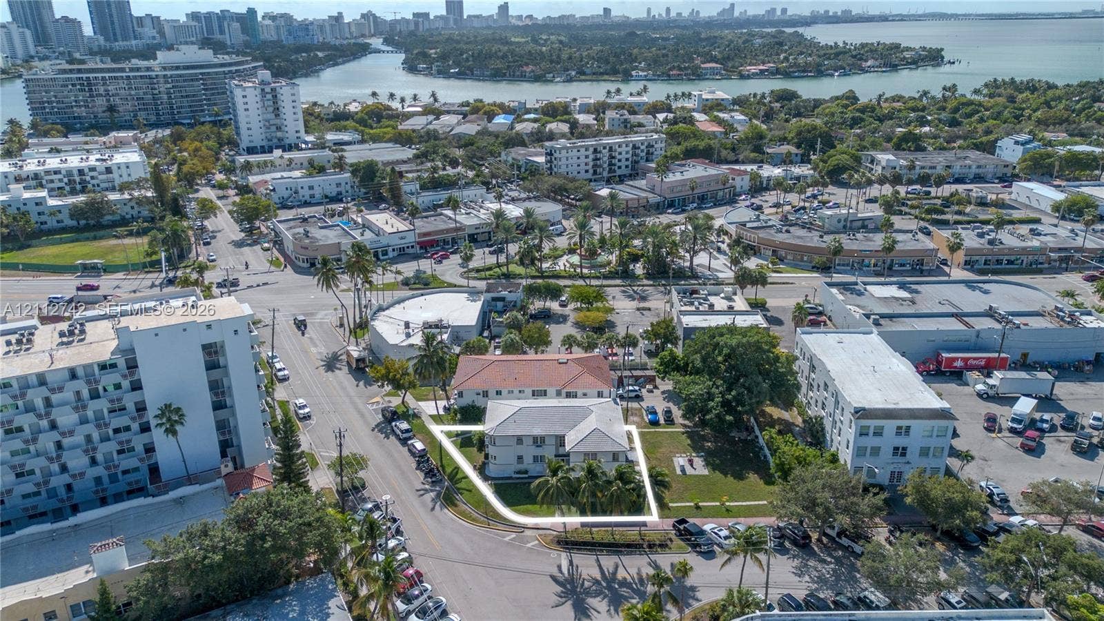 7124 Bay Drive, Miami Beach, FL 33141 Aerial perspective featuring a multi-story building with a light-toned exterior, a hipped roof, on-site surface parking, and manicured green space