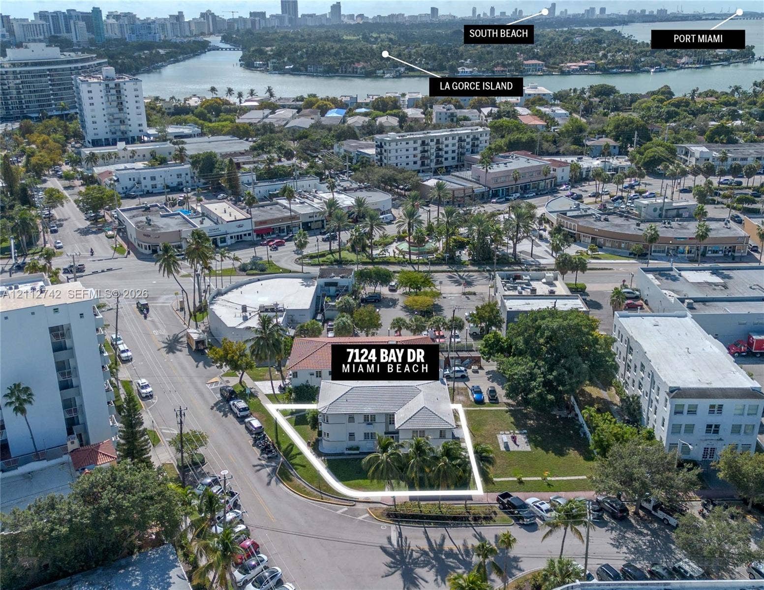 7124 Bay Drive, Miami Beach, FL 33141 Aerial perspective of a multi-unit property featuring a corner lot location, light-toned exterior finish, gabled roofline, mature palm trees, and perimeter lawn area