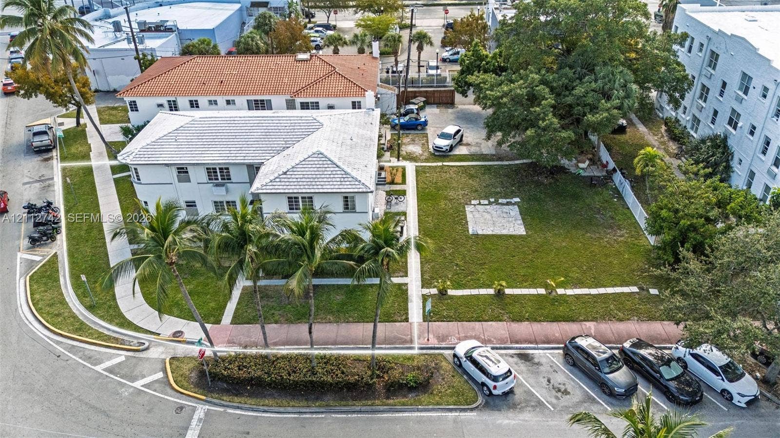 7124 Bay Drive, Miami Beach, FL 33141 Corner-lot property featuring a multi-story exterior with a light-colored finish, tiled roof, mature palm trees, expansive lawn space, and concrete walkways