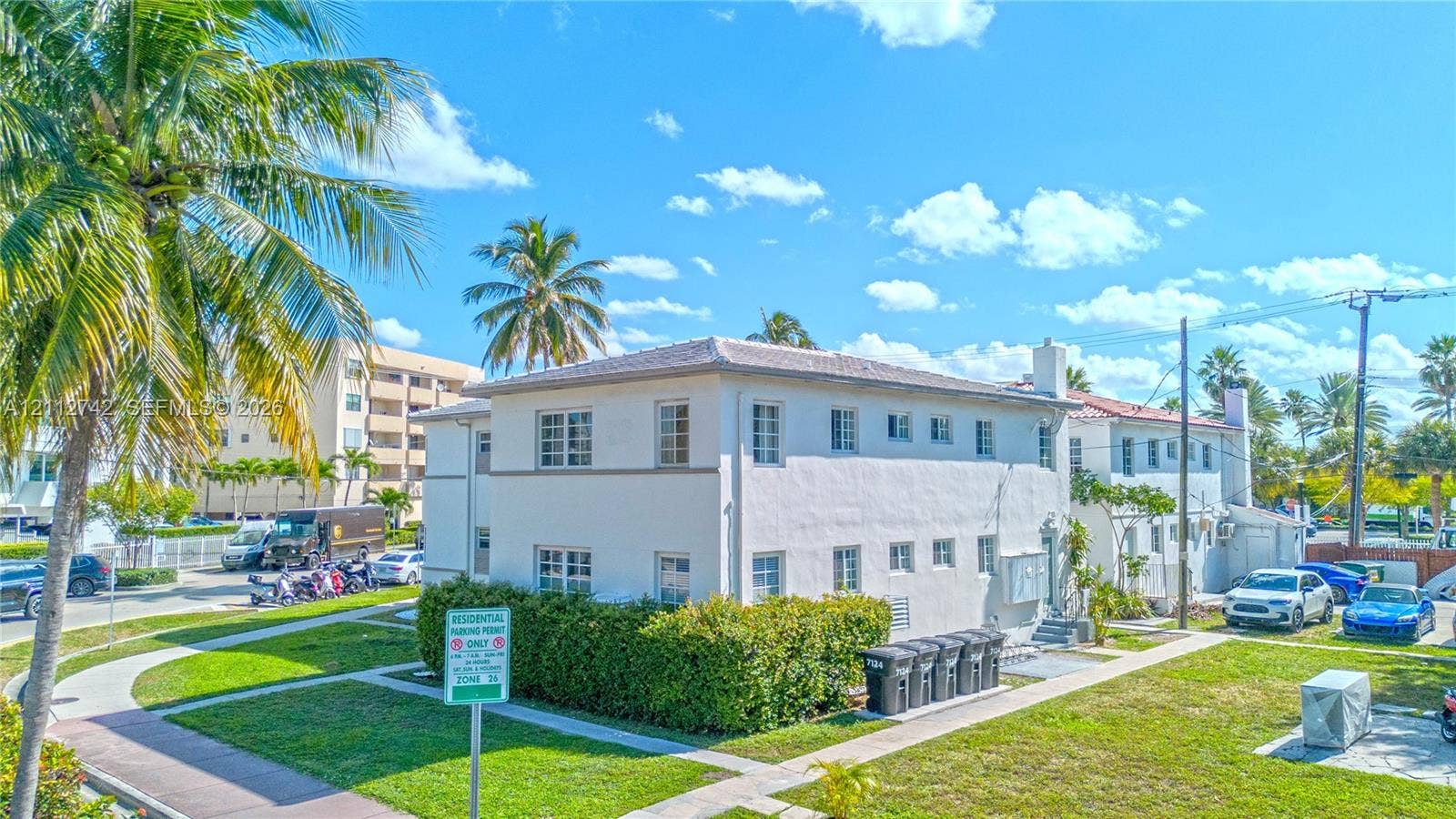 7124 Bay Drive, Miami Beach, FL 33141 Two-story exterior featuring a light-toned stucco facade, a low-pitched roof, multi-pane windows, and surrounding manicured green space