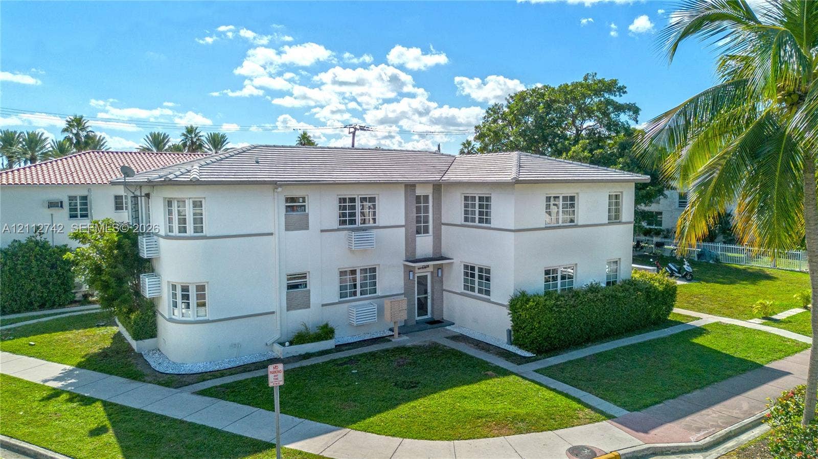 7124 Bay Drive, Miami Beach, FL 33141 Two-story exterior featuring a smooth stucco facade, hip roof with charcoal-toned shingles, rounded corner architectural detail, multi-pane windows, and concrete walkways