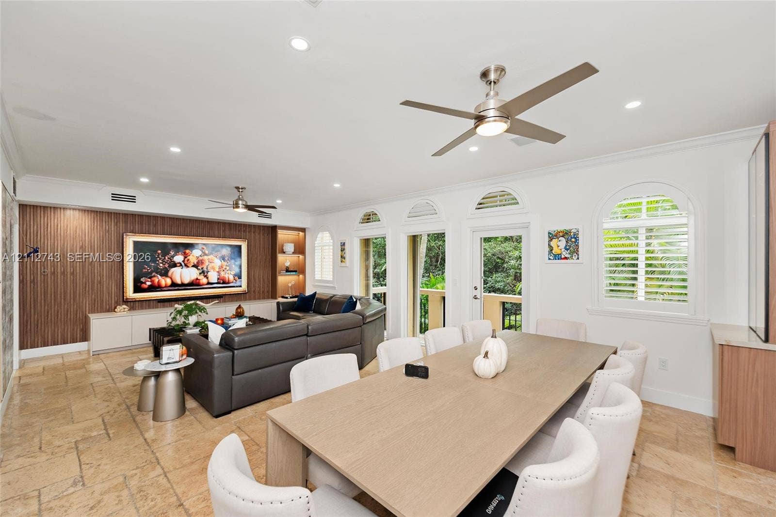 6157 Paradise Point Drive, Palmetto Bay, FL 33157 Living area featuring tile flooring, wood-finish wall paneling with integrated shelving, recessed lighting, ceiling fans, and arched windows with plantation shutters