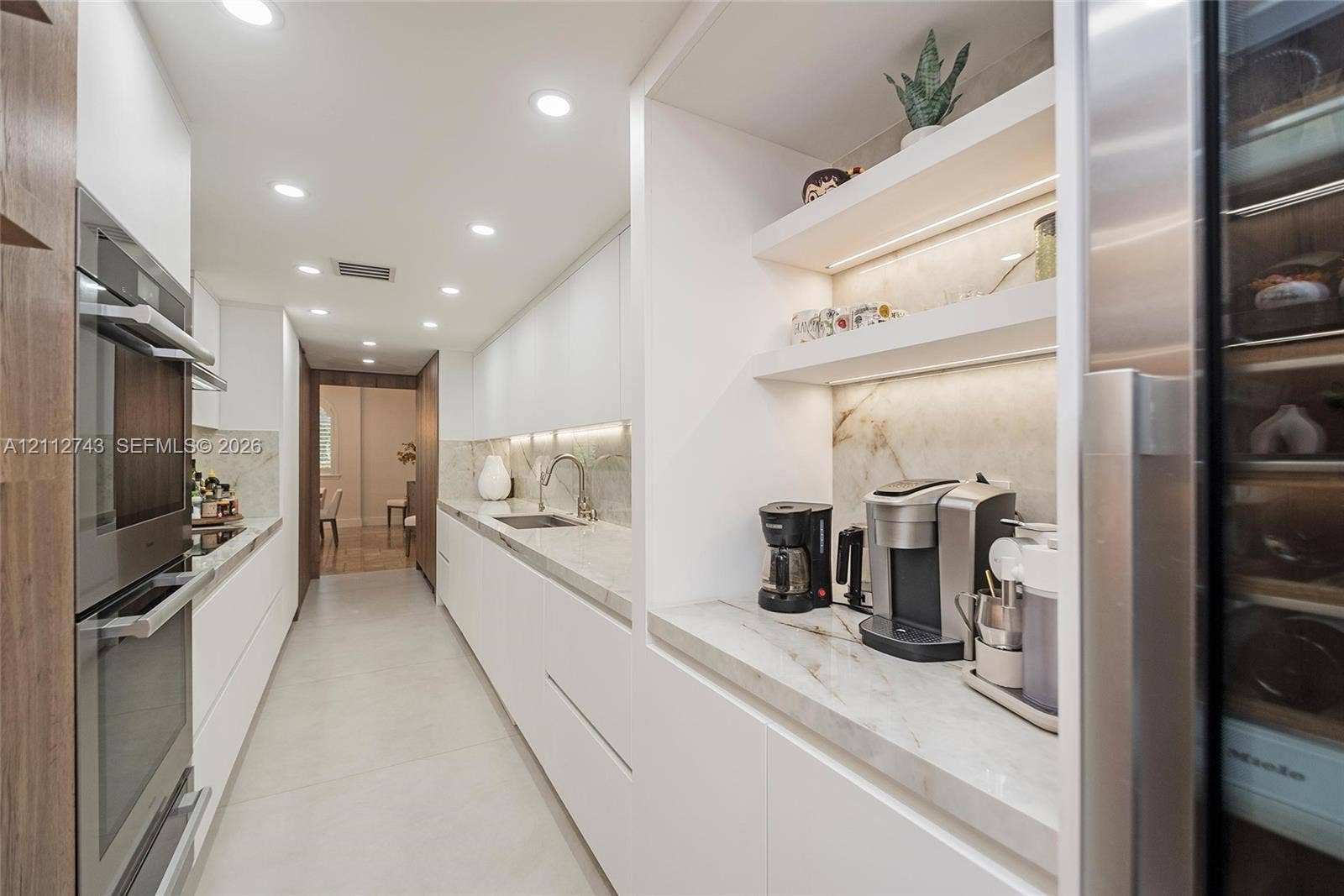 6157 Paradise Point Drive, Palmetto Bay, FL 33157 Sleek kitchen featuring flat-panel cabinetry, stone countertops, integrated stainless steel double wall ovens, built-in recessed shelving with under-cabinet lighting, and a stainless steel wine refrigerator