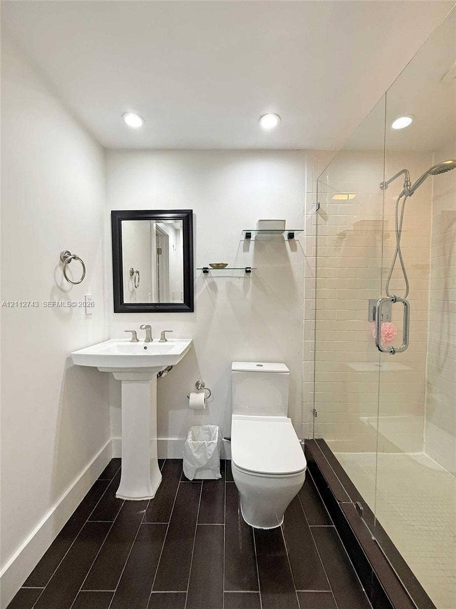 6157 Paradise Point Drive, Palmetto Bay, FL 33157 Pedestal sink with chrome widespread faucet, walk-in shower featuring a glass enclosure and wall-mounted hand shower, dark tile flooring, and integrated glass shelving