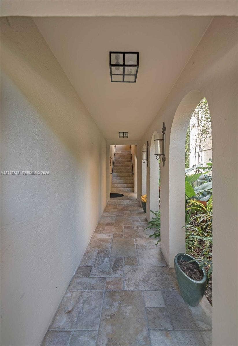 6157 Paradise Point Drive, Palmetto Bay, FL 33157 Covered walkway featuring a series of architectural arches, stone tile flooring, wall-mounted lantern-style lighting, and flush-mount overhead lighting
