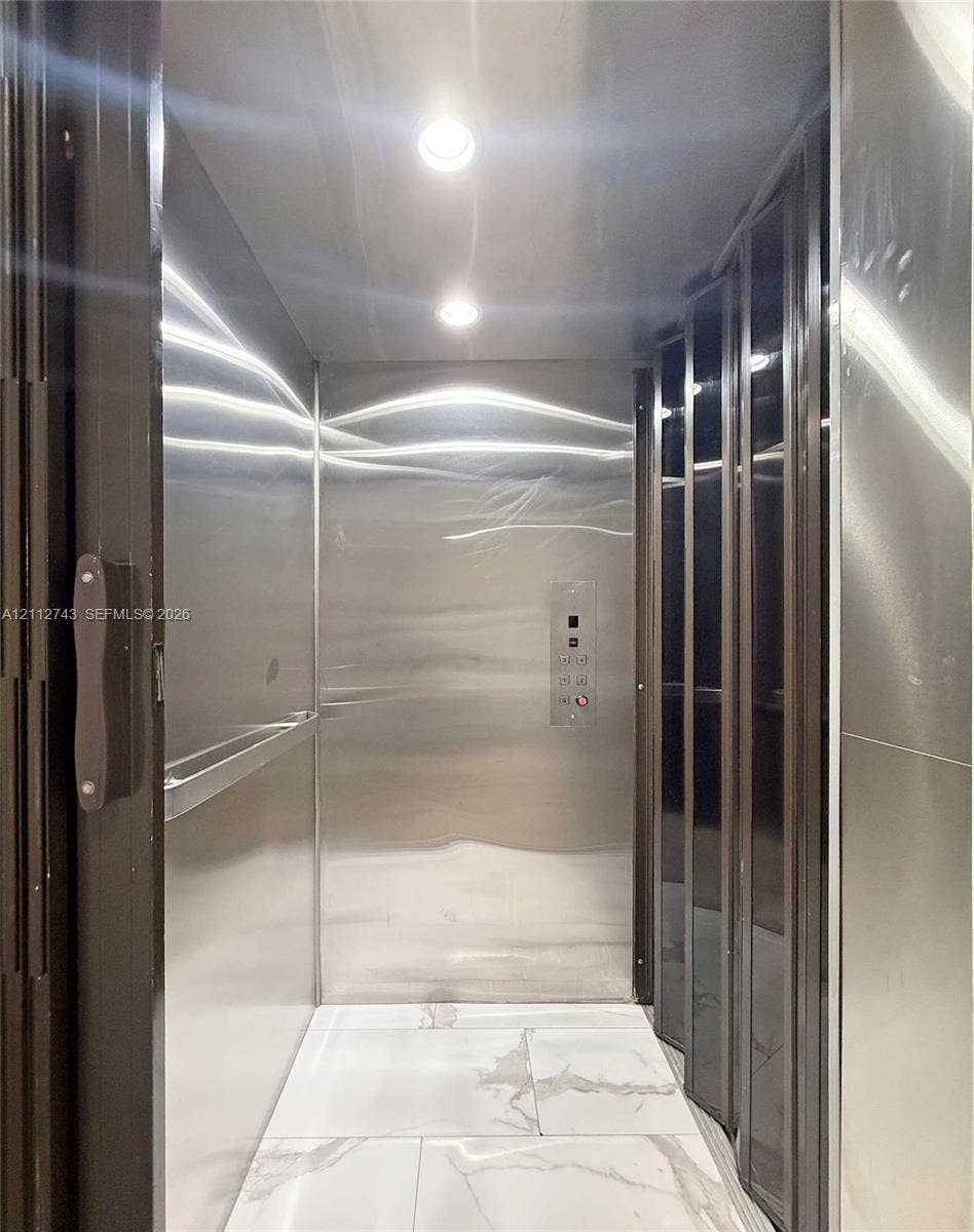 6157 Paradise Point Drive, Palmetto Bay, FL 33157 Private elevator featuring polished stainless steel wall panels, recessed ceiling lighting, a horizontal handrail, and large-format tile flooring with a veined pattern