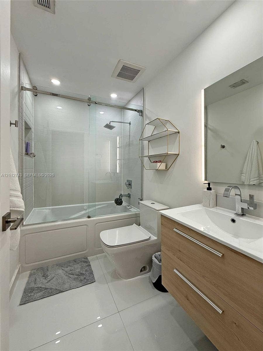 6157 Paradise Point Drive, Palmetto Bay, FL 33157 Bathroom featuring a wood-finish floating vanity with a white integrated sink, glass-enclosed shower and tub combination with a rain-style showerhead, recessed wall niche, and large-format tile flooring