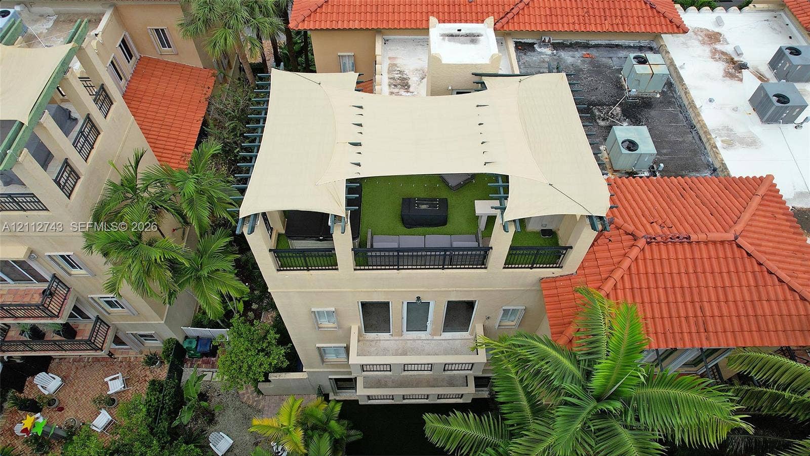 6157 Paradise Point Drive, Palmetto Bay, FL 33157 Rooftop terrace featuring synthetic turf, metal railings, and a tensile shade canopy, complemented by multiple lower-level balconies and terracotta roof tiles