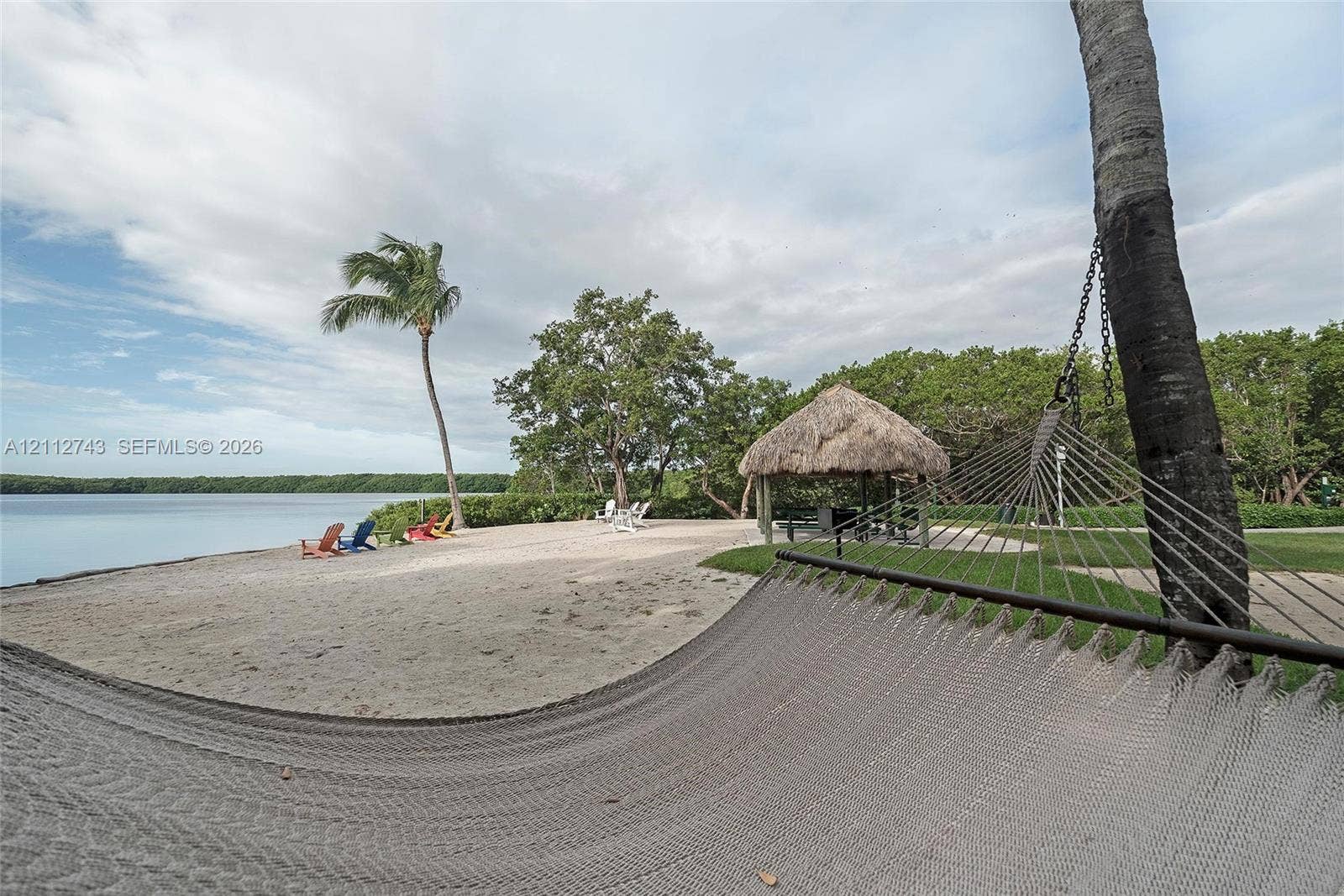 6157 Paradise Point Drive, Palmetto Bay, FL 33157 Waterfront lot featuring a sandy beach, a thatched-roof tiki hut, mature palm trees, and lush perimeter landscaping