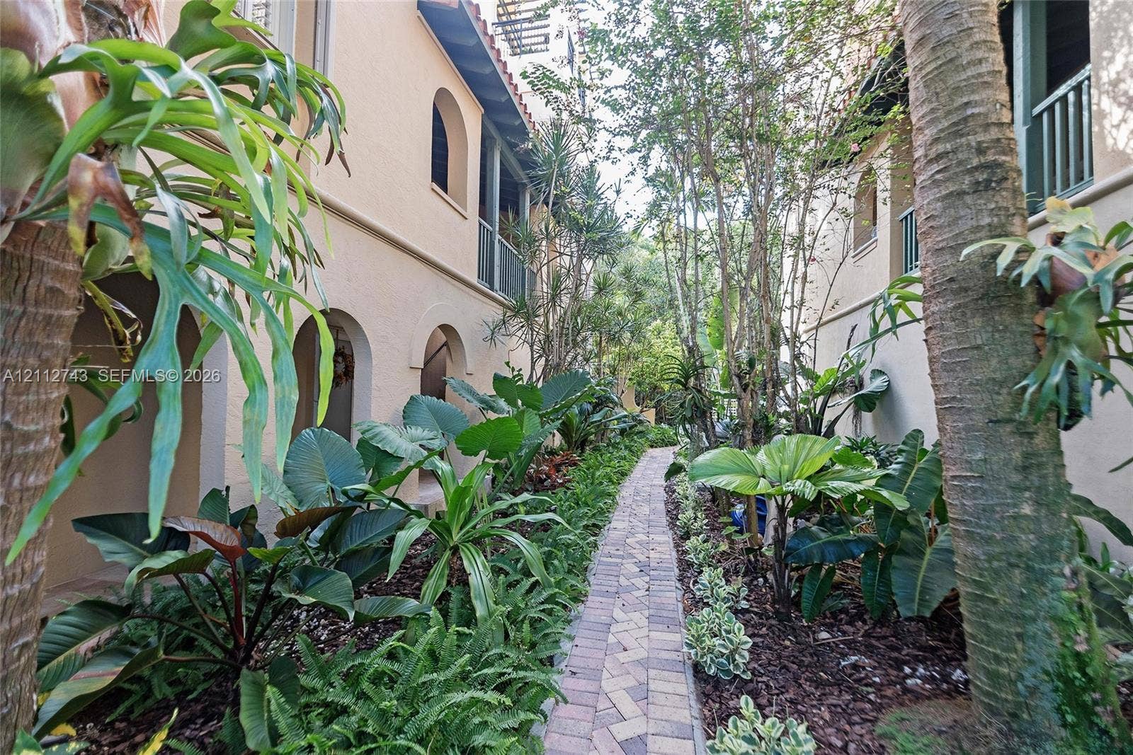 6157 Paradise Point Drive, Palmetto Bay, FL 33157 Stucco exterior featuring arched breezeways and second-story balcony railings, accented by a herringbone-patterned paver walkway and mature landscaping