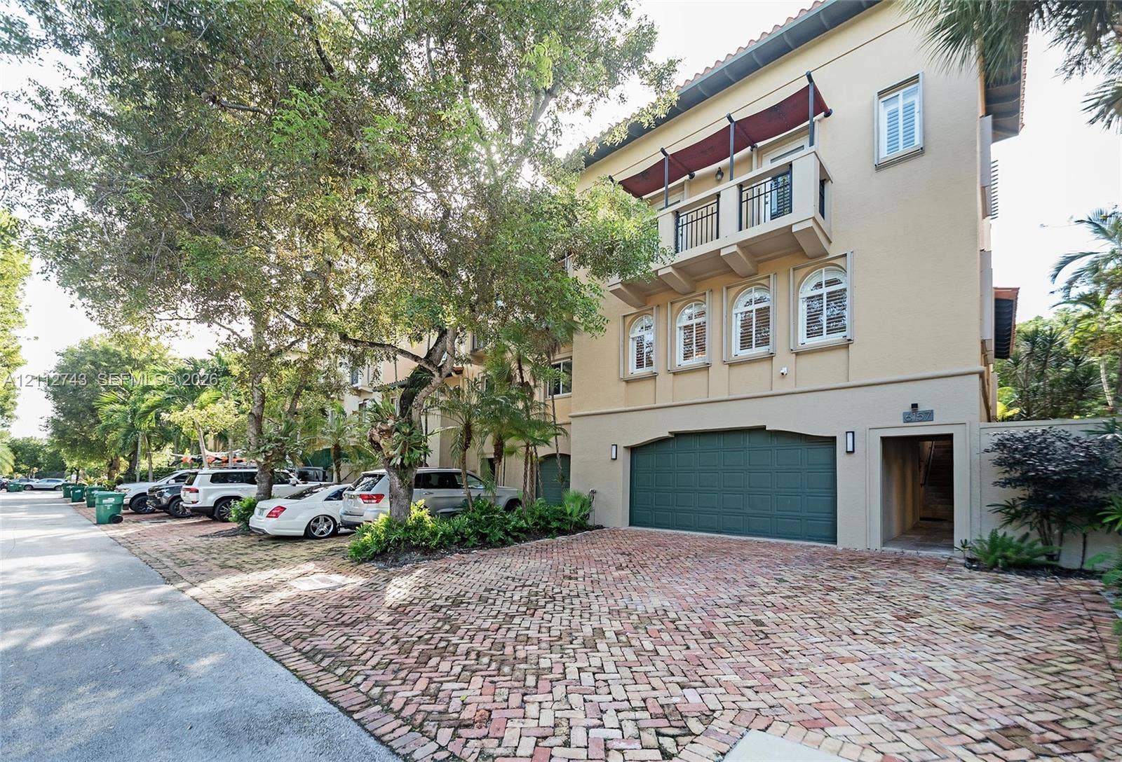 6157 Paradise Point Drive, Palmetto Bay, FL 33157 Three-story exterior featuring a brick-paved driveway, multi-car garage with arched detailing, balcony with metal railing, and arched window accents