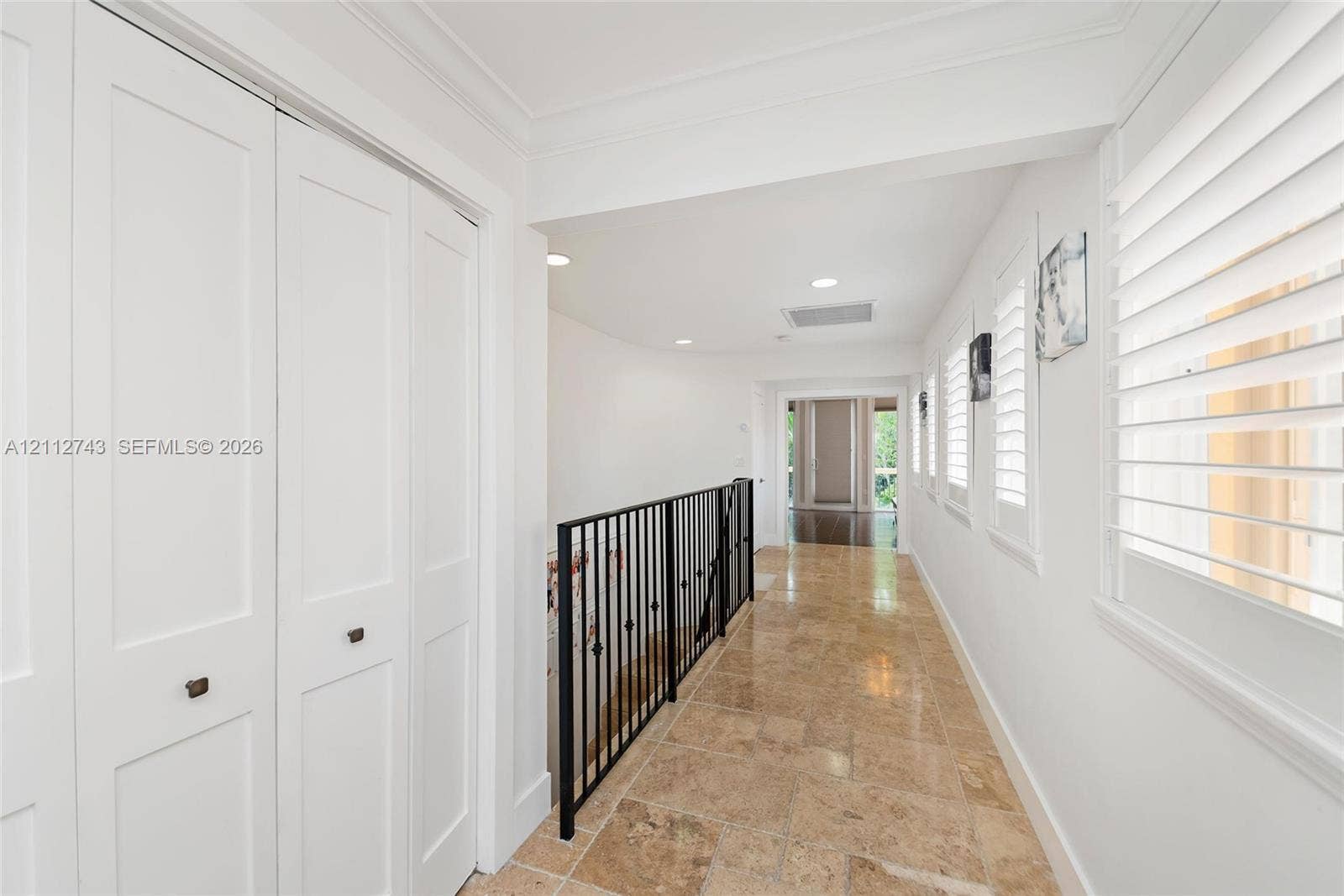 6157 Paradise Point Drive, Palmetto Bay, FL 33157 Hallway featuring stone tile flooring, white bi-fold closet doors, recessed lighting, plantation shutters, and a wrought-iron railing