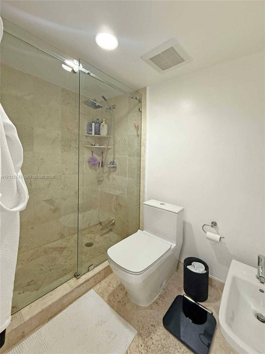 6157 Paradise Point Drive, Palmetto Bay, FL 33157 Walk-in shower featuring a frameless glass enclosure, stone tile surround, and a handheld shower fixture, complemented by natural stone flooring and a high-profile toilet