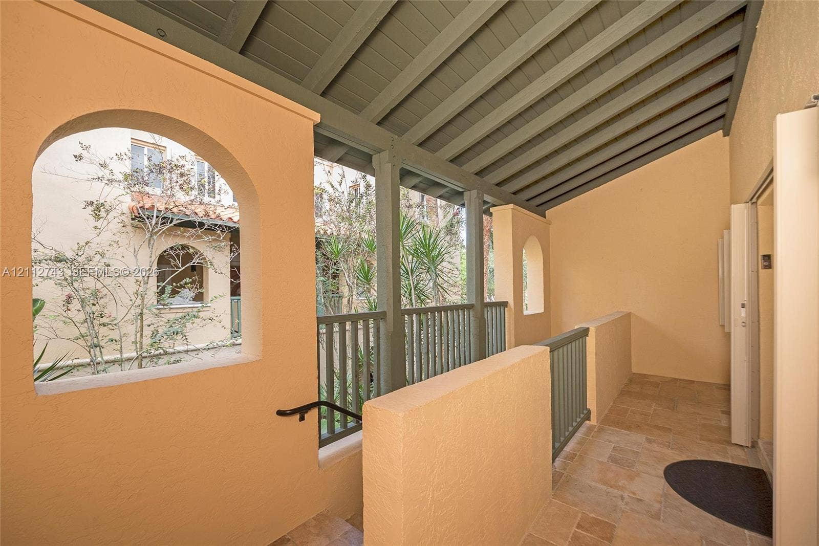 6157 Paradise Point Drive, Palmetto Bay, FL 33157 Covered exterior walkway featuring arched openings, stone-tile flooring, painted wood-beam ceiling, and vertical slat railings