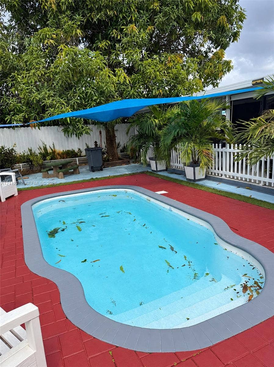 1210 N 71st Avenue, Hollywood, FL 33024 In-ground pool with molded steps, surrounded by red brick-patterned paving and a gray bullnose coping border, complemented by white picket fencing and a blue overhead tension shade sail