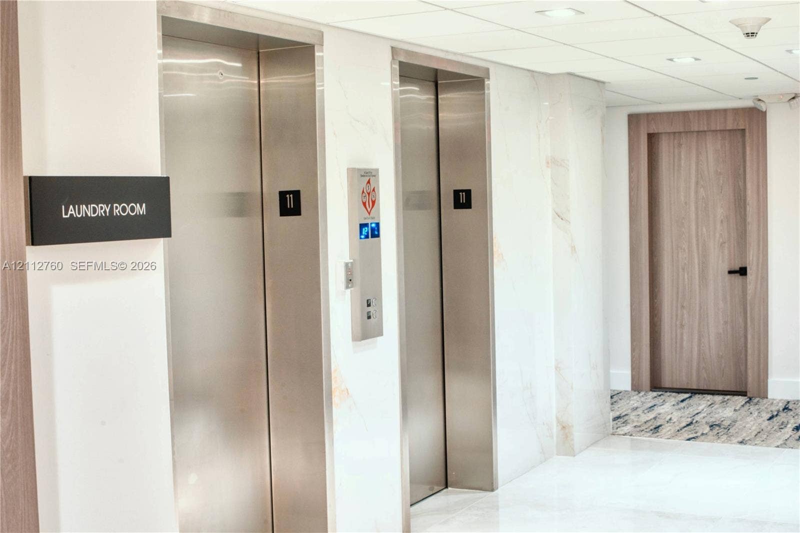 5825 Collins Avenue, #11H, Miami Beach, FL 33140 Elevator lobby featuring stainless steel doors, white stone-finish wall cladding, recessed ceiling lighting, and a wood-finish interior door