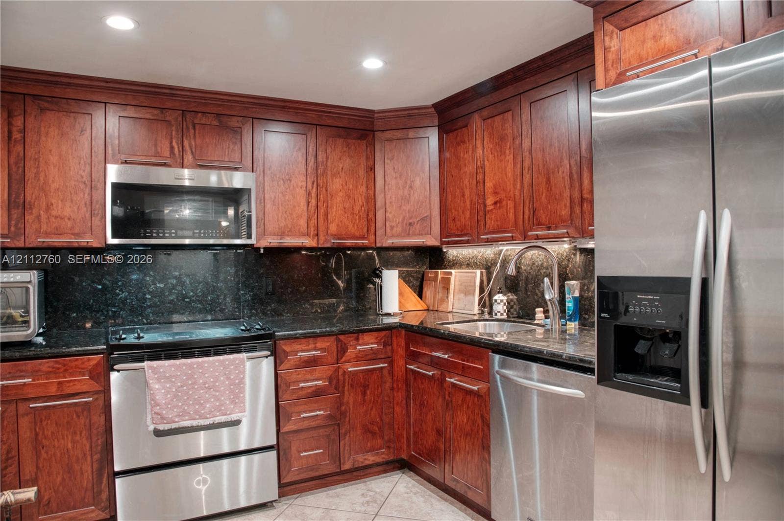 5825 Collins Avenue, #11H, Miami Beach, FL 33140 Kitchen featuring cherry-finish cabinetry, dark stone countertops, stainless steel appliances, a recessed under-cabinet lighting system, and tile flooring