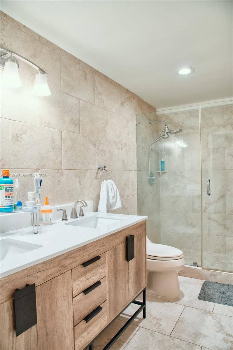 5825 Collins Avenue, #11H, Miami Beach, FL 33140 Double vanity featuring a white countertop with dual integrated sinks and a wood-finish cabinet base, complemented by a walk-in glass-enclosed shower with neutral tile surround