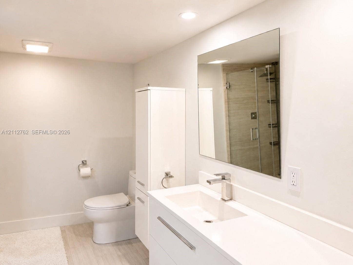White floating vanity with integrated rectangular sink, sleek chrome faucet, tall built-in linen storage cabinet, wall-mounted mirror, and glass-enclosed shower