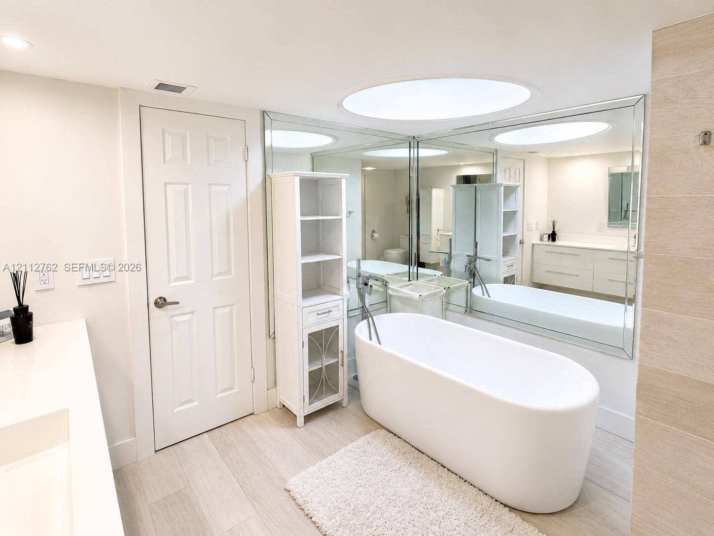 Freestanding soaking tub with a floor-mounted faucet, wall-to-wall vanity mirror, neutral-toned flooring, and built-in cabinetry
