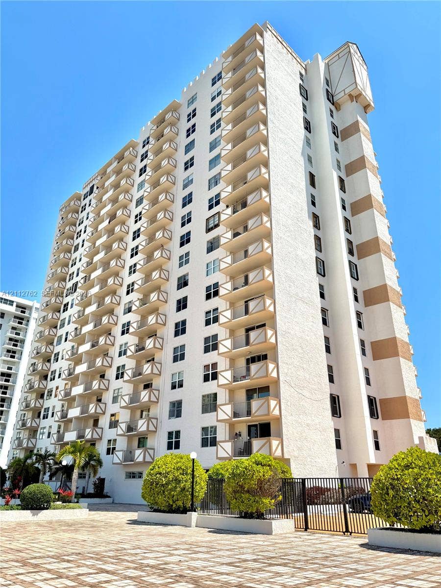 High-rise building exterior featuring tiered balconies with railing accents, masonry facade, and paved ground-level hardscaping