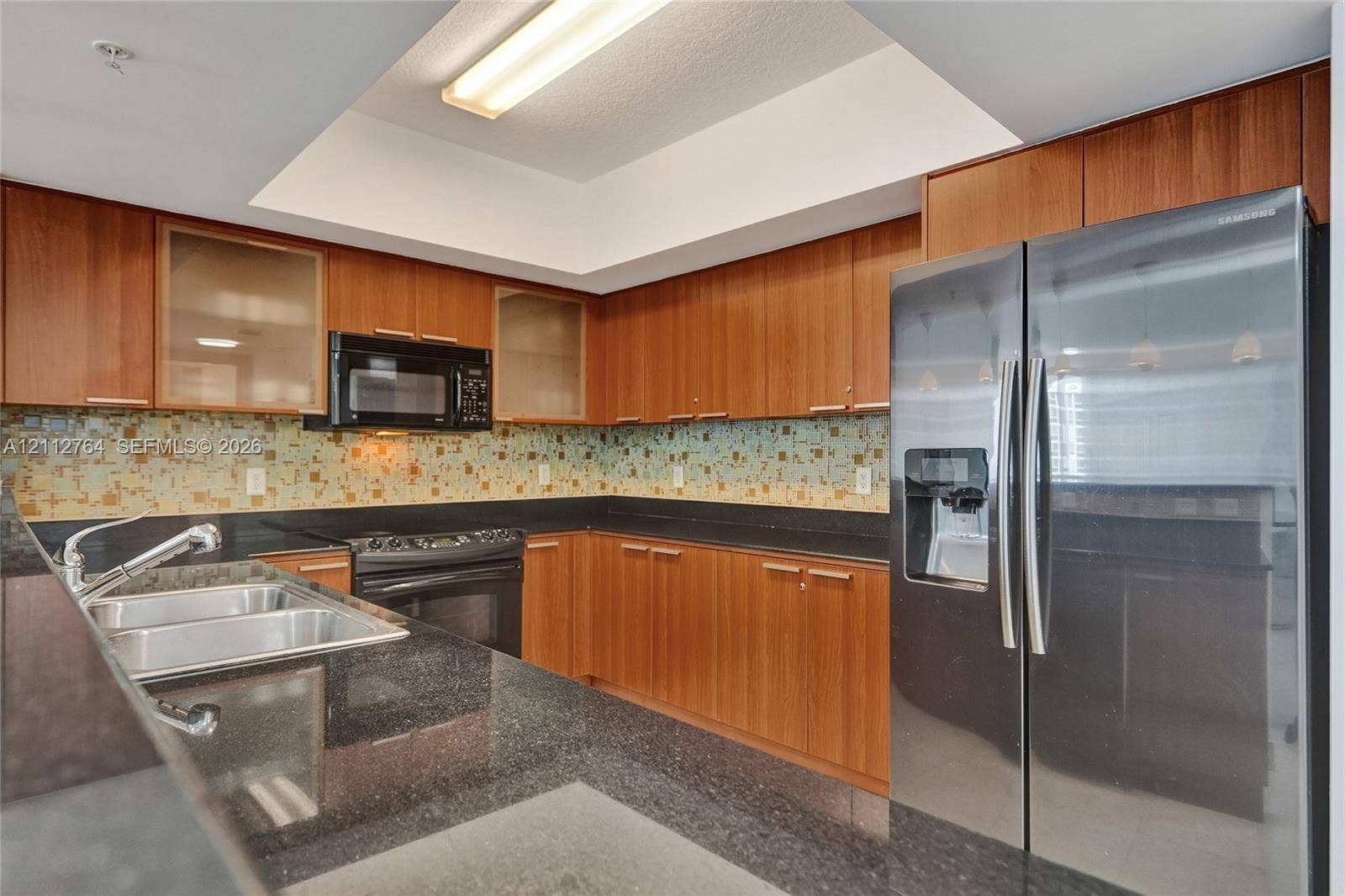Kitchen featuring wood cabinetry, dark stone-finish countertops, multi-toned glass tile backsplash, recessed tray ceiling, and stainless steel appliances