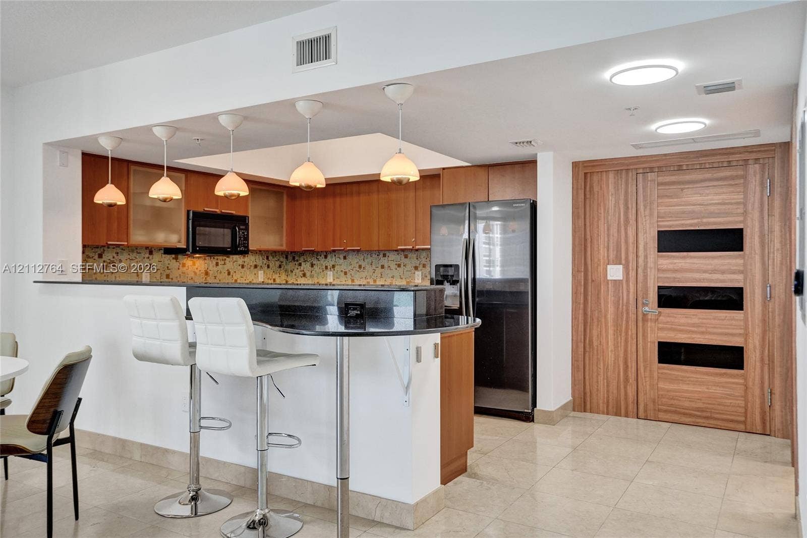 Kitchen featuring wood-finish cabinetry, stone countertop breakfast bar, mosaic tile backsplash, pendant lighting, and full-height wood-finish door with glass accents
