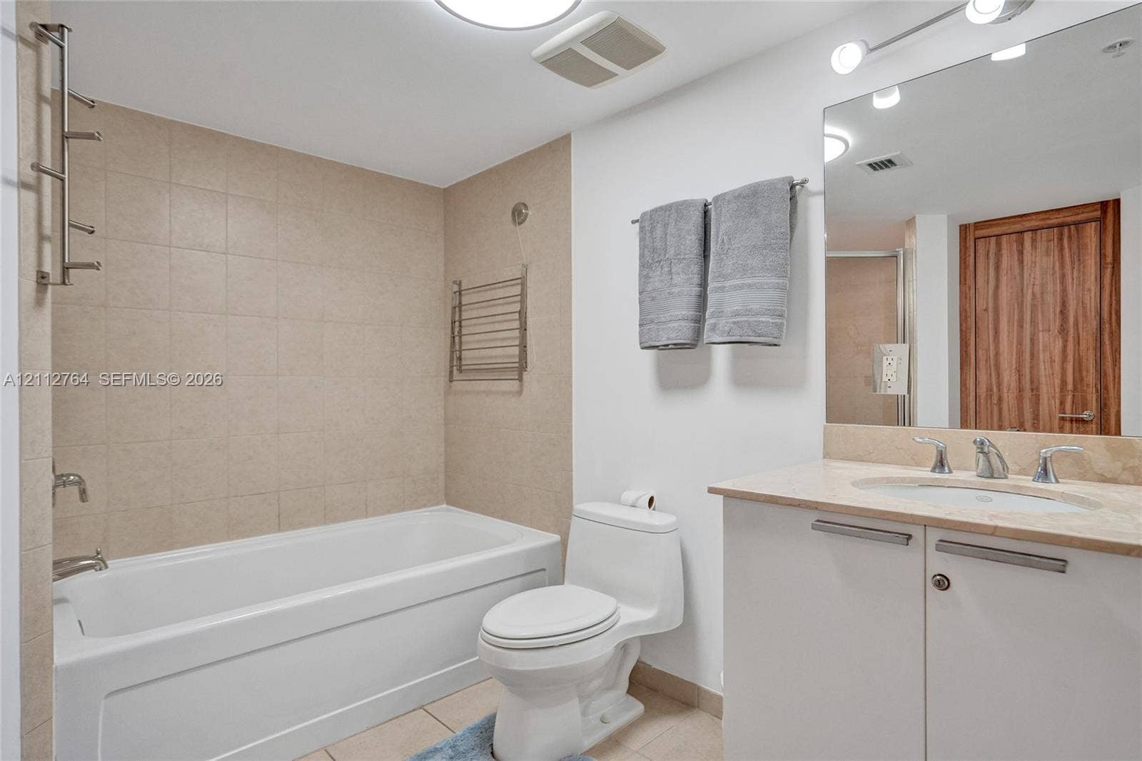 Bathroom featuring a deep soaking tub with neutral tile surround, vanity with stone-finish countertop, undermount sink, and full-width vanity mirror with integrated lighting