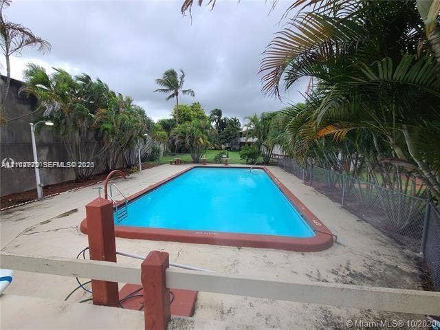 Rectangular in-ground swimming pool featuring a terra cotta border, metal handrail, concrete pool deck, perimeter fencing, and mature palm trees