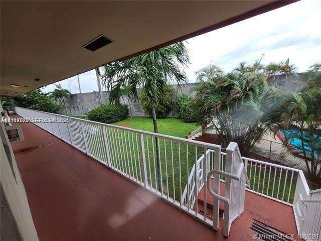 Covered balcony featuring white metal railings, stairs, concrete flooring, exterior lighting, and overlooking a green space with a swimming pool and mature palm trees