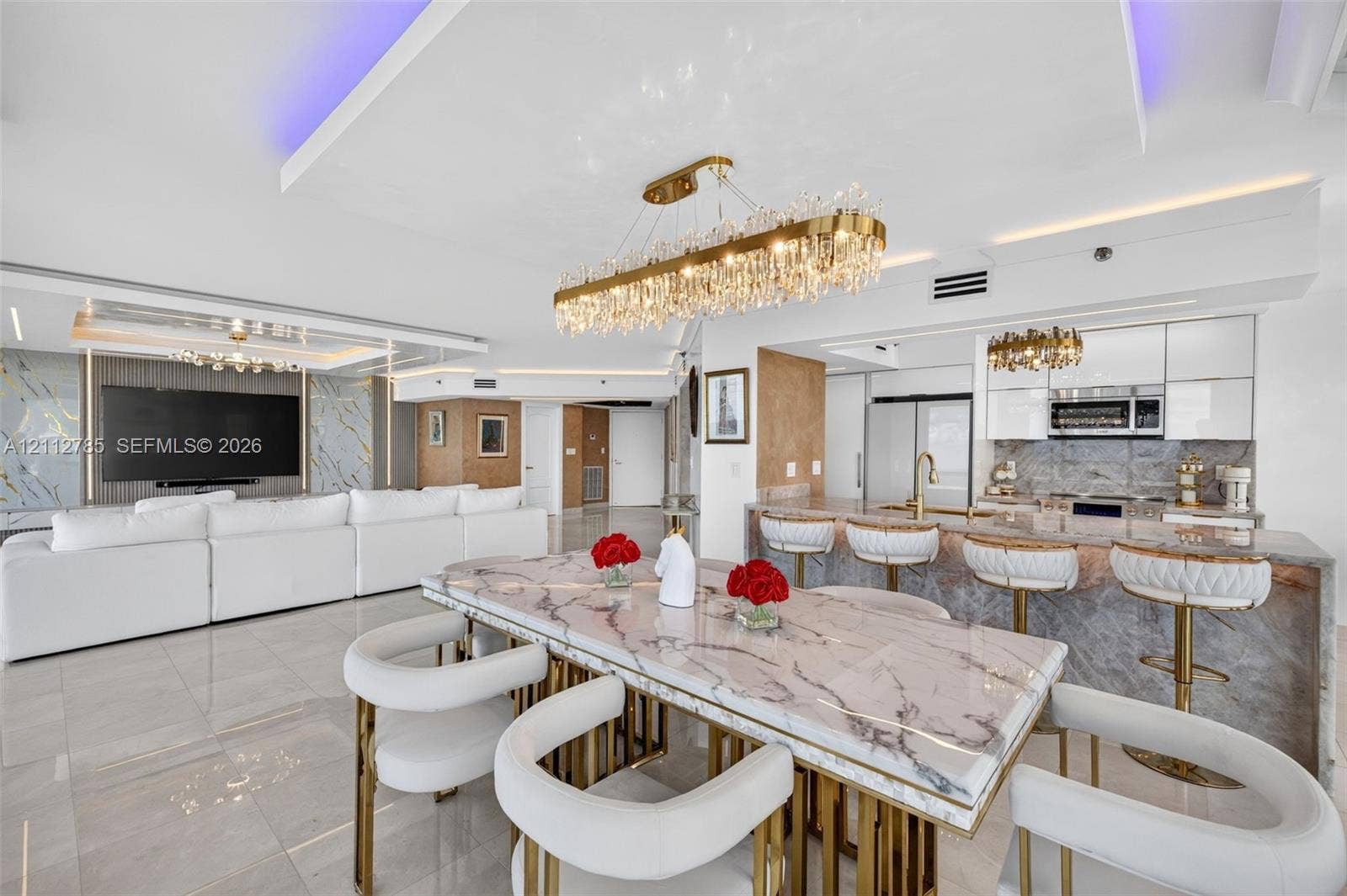 16425 Collins Avenue, #2016, Sunny Isles Beach, FL 33160 Open-concept living space featuring high-gloss tile flooring, stone-surfaced island and dining table, gold-toned crystal chandeliers, and recessed tray ceilings with integrated ambient lighting