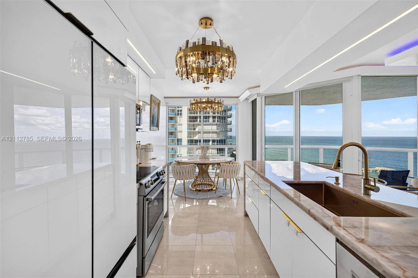 16425 Collins Avenue, #2016, Sunny Isles Beach, FL 33160 Contemporary kitchen featuring high-gloss white cabinetry, stone-surface countertops, integrated stainless steel appliances, tiered crystal chandeliers, and floor-to-ceiling windows with coastal scenery