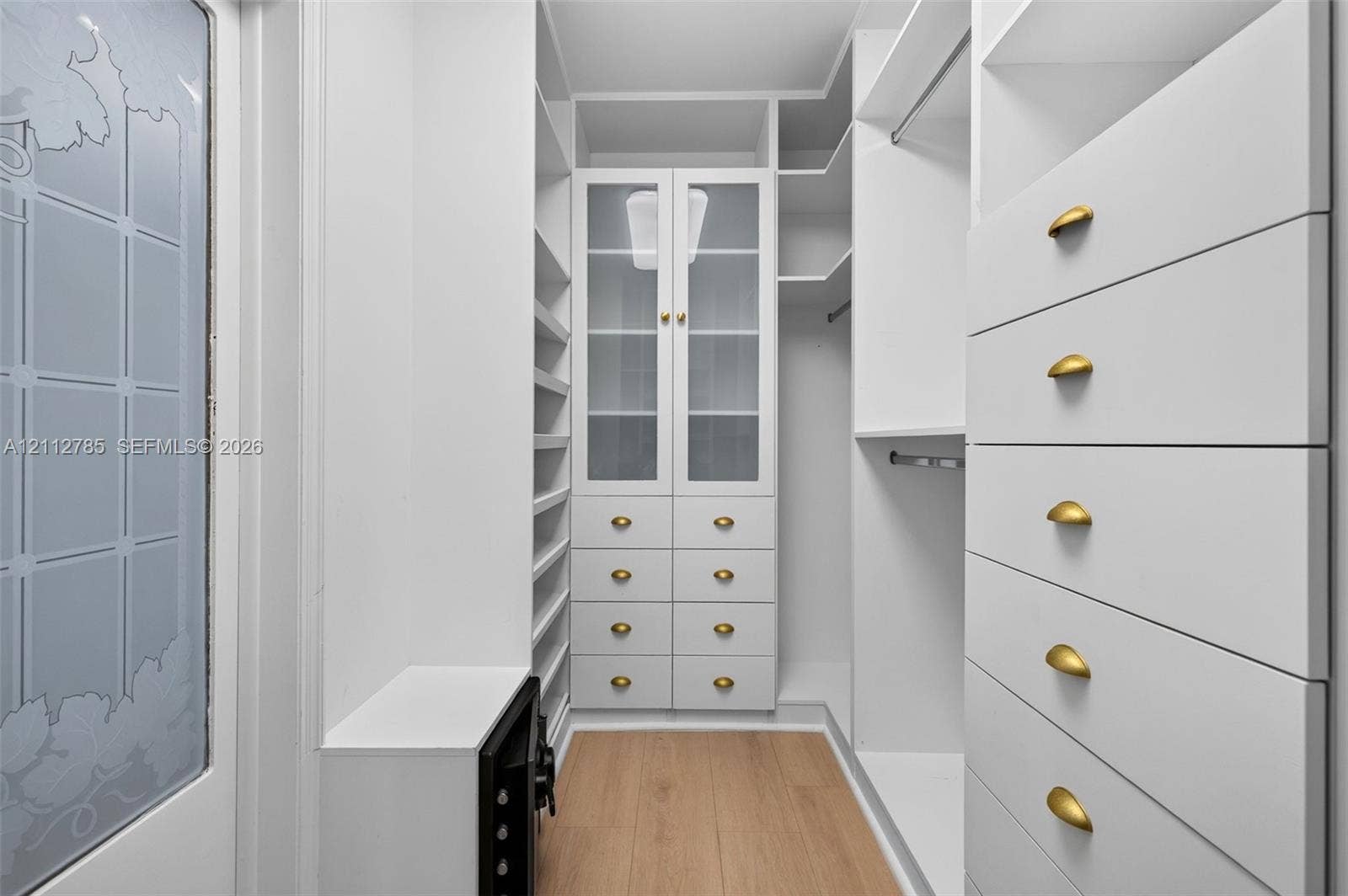 16425 Collins Avenue, #2016, Sunny Isles Beach, FL 33160 Custom walk-in closet featuring white built-in cabinetry, glass-front upper storage, soft-gold tone hardware, integrated drawer banks, and wood-finish flooring