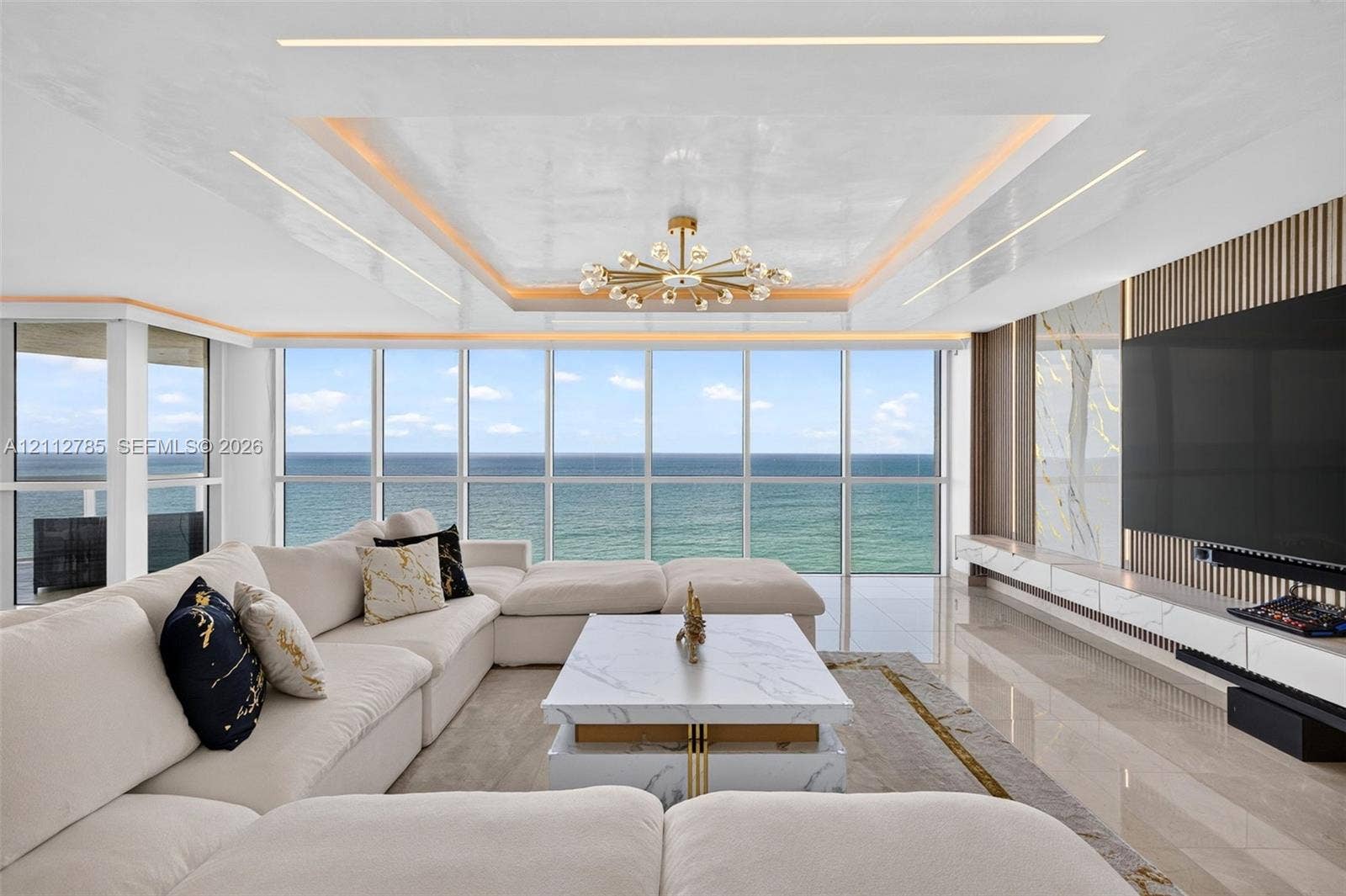 16425 Collins Avenue, #2016, Sunny Isles Beach, FL 33160 Floor-to-ceiling windows featuring ocean scenery, tray ceiling with recessed cove lighting, central sputnik-style chandelier, and custom built-in media wall with stone cladding and vertical wood slat detailing